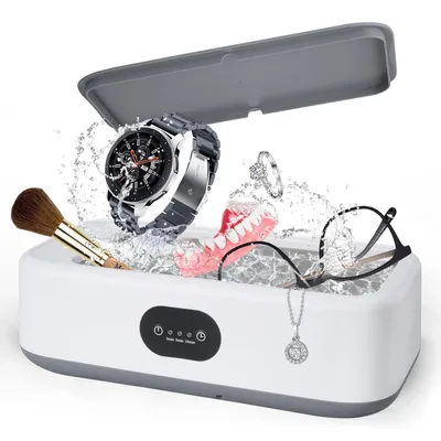 A white ultrasonic cleaner with a gray lid open, filled with water and various items being cleaned, including a watch, glasses, a necklace, a makeup brush, and dentures. Water is splashing around the items.