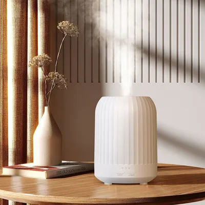 A white ultrasonic diffuser emitting mist sits on a wooden table next to a vase with dried flowers and a book.