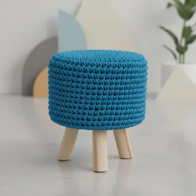 A blue crocheted stool with wooden legs on a light-colored floor, with abstract shapes in the background.