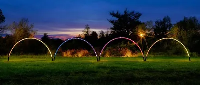 Four illuminated arches are set up in a grassy field at dusk.