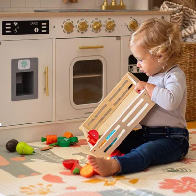 A toddler plays with a wooden toy kitchen set and play food on a mat.
