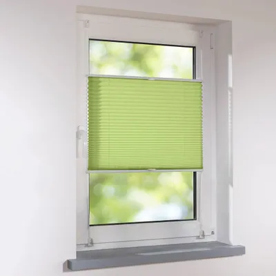 A green pleated blind is installed in a white window frame, with blurred green foliage visible through the window.