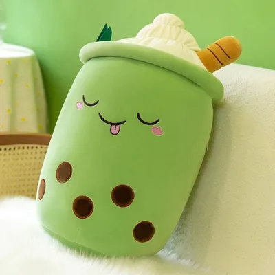 A plush toy shaped like a green bubble tea drink with a smiling face is resting on a white furry surface, with a green background and a hint of a table with a patterned cloth in the background.