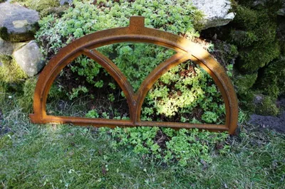 A rusty metal window frame is placed on the ground in a garden setting, surrounded by greenery and rocks.