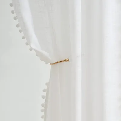 A close-up of a white sheer curtain with pom-pom trim, held by a gold curtain tie.
