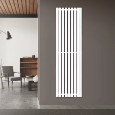 A white vertical radiator is mounted on a gray wall in a modern room with a blurred background showing furniture and a lamp.