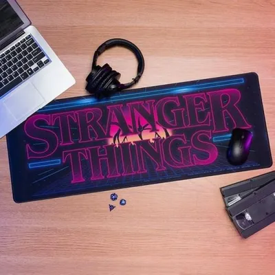 A desk setup with a laptop, headphones, a Stranger Things themed mousepad, a computer mouse, and a VHS tape.