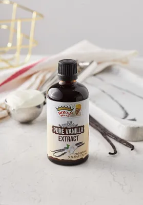 A bottle of vanilla extract is placed on a white surface with some kitchen utensils and vanilla beans in the background.