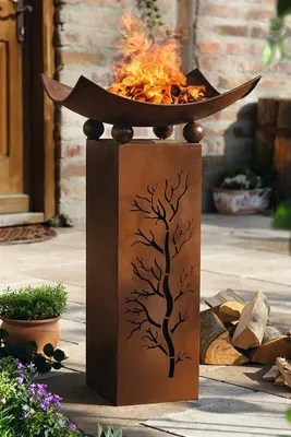 A metal fire pit with a tree design on a pedestal, set on a patio with firewood and plants.