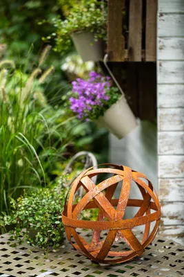 A decorative metal sphere sits on a patterned surface in a garden setting, with plants and hanging baskets in the background.