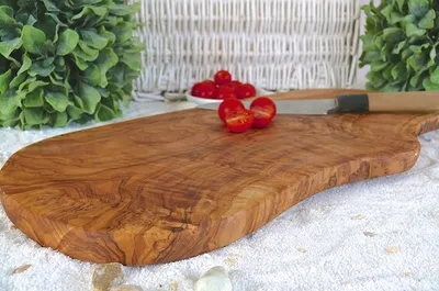 A rustic olive wood cutting board with cherry tomatoes and a knife on a white textured surface, with greenery in the background.