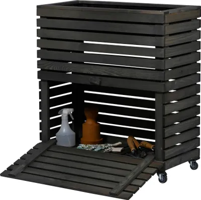 A dark gray wooden raised garden bed with storage space underneath, shown outdoors with gardening tools and a spray bottle inside.