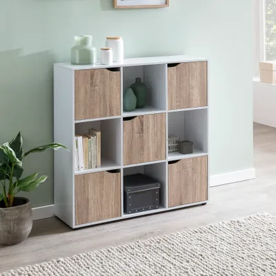 A white and wood-tone cube shelf unit with various items on and in its shelves, placed in a room with a light green wall and a rug in the foreground.
