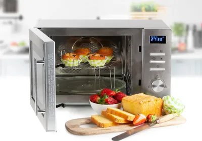 A stainless steel microwave oven with its door open, showing muffins inside. In front of the microwave are strawberries, sliced bread, and a loaf of cake on a wooden cutting board.