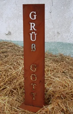 A weathered metal sign with the text "Grüß Gott" stands upright in a pile of straw, with a textured wall in the background.