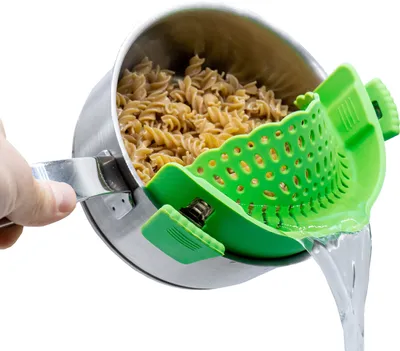 A hand is holding a pot with pasta and a green silicone strainer attached, while water is being drained.