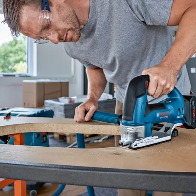 A man is using a blue and black jigsaw to cut a curved edge on a wooden board in a workshop setting.