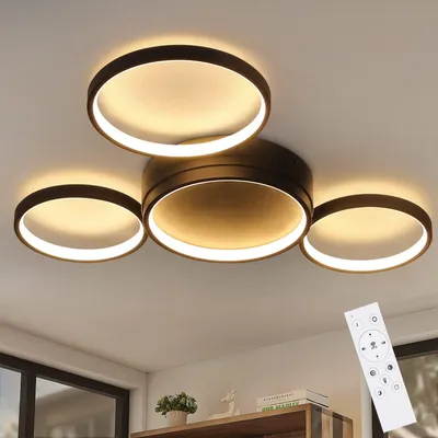 A modern black LED ceiling light with four rings is shown on a white ceiling. A remote control is visible in the bottom right corner.