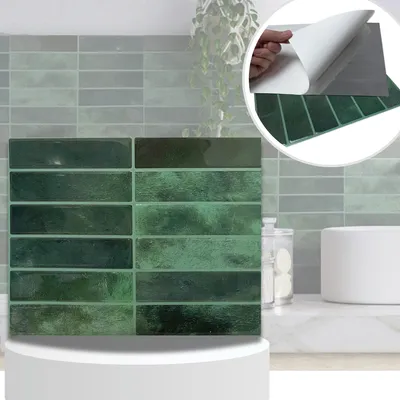 A close-up view of green peel-and-stick wall tiles, with a hand peeling the backing off a sample tile in the upper right corner. The tiles are arranged in a brick pattern on a countertop, with a blurred background of a kitchen or bathroom.