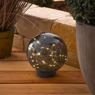 A solar-powered garden sphere with LED lights sits on a wooden deck, surrounded by plants and a stone wall.