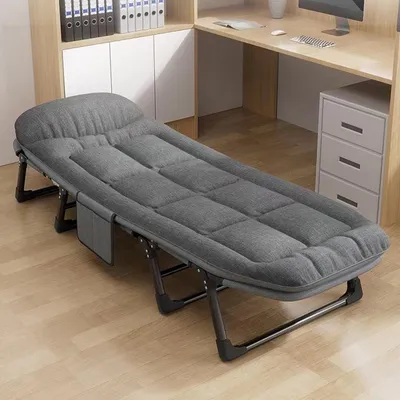 A gray folding cot is placed on a wooden floor next to a desk and shelves.