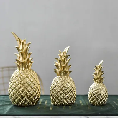 Three golden pineapple ornaments of varying sizes are displayed on a dark green cloth.