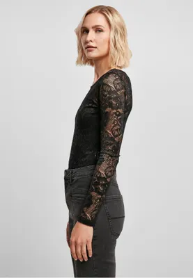 A woman is shown from the side wearing a black lace long-sleeved bodysuit and dark wash jeans against a plain gray background.