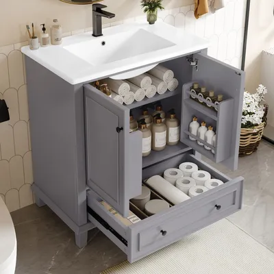 A grey bathroom vanity with a white ceramic sink, open doors revealing storage, and a partially open drawer filled with toiletries.
