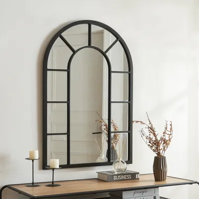A black arched mirror with a grid design is mounted on a white wall above a wooden console table.