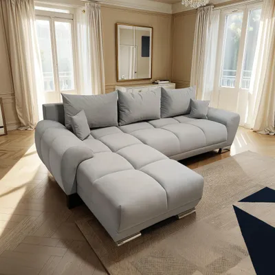 A light gray sectional sofa is placed in a well-lit living room with large windows and sheer curtains.