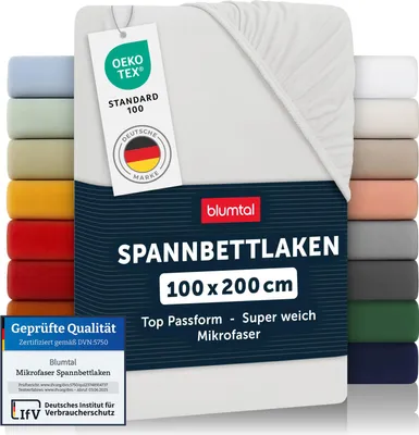 A white fitted sheet is displayed prominently, with a stack of sheets in various colors visible on the left and right sides. Several tags and certifications are attached to the sheet.