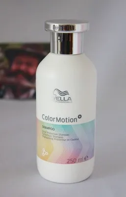 A white bottle of Wella Professionals ColorMotion+ shampoo with a silver cap is shown against a light gray and white background. A blurred image of a person is visible in the background.
