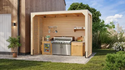 An outdoor kitchen shelter with a grill, refrigerator, and storage cabinets, set in a backyard with greenery and a wooden building.