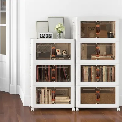 Two stacks of white storage boxes with clear doors, filled with books and other items, are placed against a white wall on a wooden floor.
