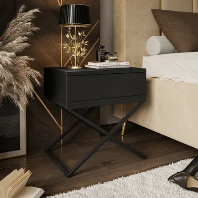 A black nightstand with a drawer is placed next to a bed in a bedroom setting. The nightstand has a gold-colored, X-shaped base. A lamp with a black pleated shade and a gold stem shaped like branches sits on top of the nightstand, along with a stack of boo