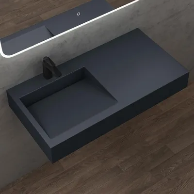 A modern, matte dark gray washbasin with a black faucet is mounted on a wall above a wooden floor. A backlit mirror is above the washbasin.