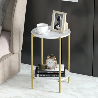 A small round side table with a marble-look top and gold legs, placed next to a bed. It holds a cup, a framed picture, and books.