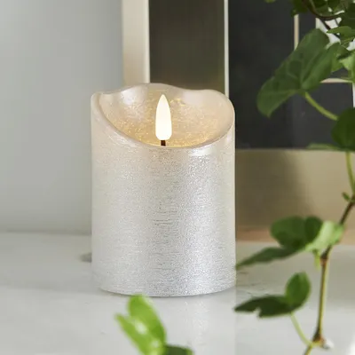 A close-up of a silver LED candle with a flickering flame, placed on a white surface with greenery in the foreground and background.