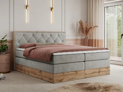 A bedroom scene featuring a king-size boxspring bed with a tufted headboard, dressed with bedding and decorative pillows. The bed has a wooden base and is placed in a room with soft lighting and decorative elements.