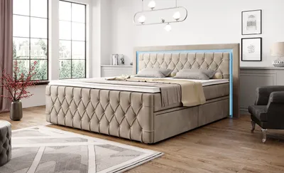 A bedroom scene with a large bed as the focal point, decorated with pillows and throws. The room has large windows, a rug, and a chair.