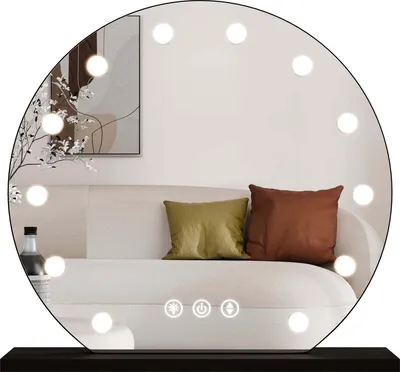 A round mirror with lights is placed on a dark surface, reflecting a sofa with pillows and a framed picture on the wall.