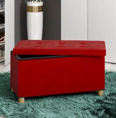 A red storage ottoman is shown in an interior setting, possibly a room or hallway, with a blurred background that includes a white vase and part of a closet or cabinet.