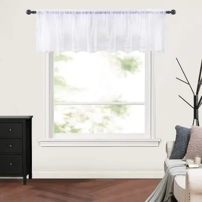 A white sheer valance is hung on a curtain rod above a window. The window looks out onto trees and greenery. To the left is a black dresser, and to the right is a sofa with pillows.
