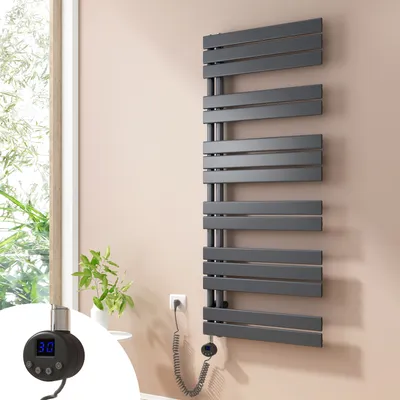 A dark gray electric towel radiator is mounted on a light pink wall next to a window with greenery visible outside. A small black digital thermostat with a blue display is attached to the radiator.