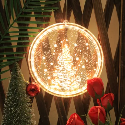 A festive Christmas decoration with a glowing tree scene hanging against a wooden fence, surrounded by greenery and red roses.