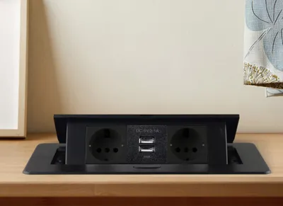 A black pop-up power outlet with two European sockets and two USB ports is shown on a wooden desk.