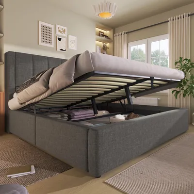 A grey upholstered bed with its storage compartment open, revealing folded linens inside. The bed is in a bedroom setting with artwork on the wall and curtains in the background.