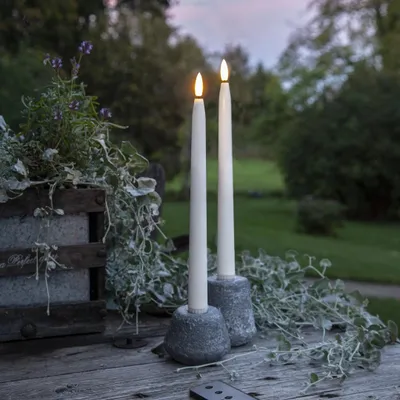 Two LED candles with flickering flames stand on a wooden table in an outdoor setting, with greenery and trees in the background.