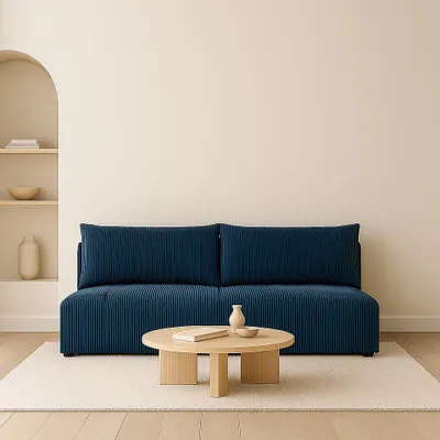 A blue corduroy sofa with two large cushions is centered in a room with a light-colored wall and a built-in arched shelf on the left. A round wooden coffee table with a book and a small vase sits in front of the sofa on a cream rug.