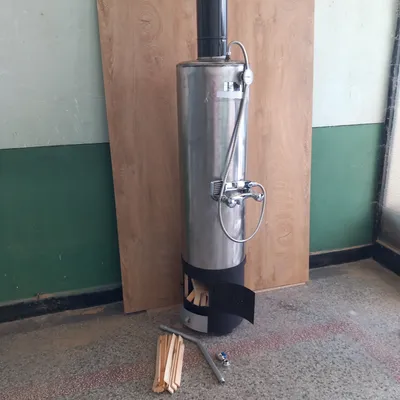 A stainless steel wood-fired water heater with a shower head and faucet is standing on a floor in front of a wooden board. A small pile of firewood is on the floor next to it.